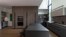 View of the island with absolute black leathered countertops and a backdrop of warm wood tones in the tower wall.